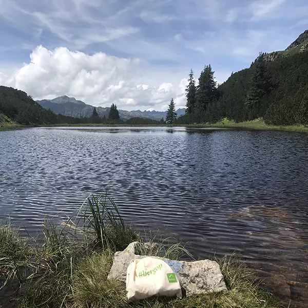 Een rustig bergmeer met helder water, omringd door bomen en bergtoppen in de verte, onder een gedeeltelijk bewolkte hemel. Op de voorgrond ligt een rots bij de waterkant, met daarop een herbruikbare tas met het opschrift „Hubergut“.